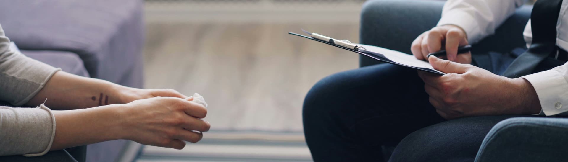 Two people seated opposite each other in a quiet room, one holding a tissue, the other making notes on a clipboard