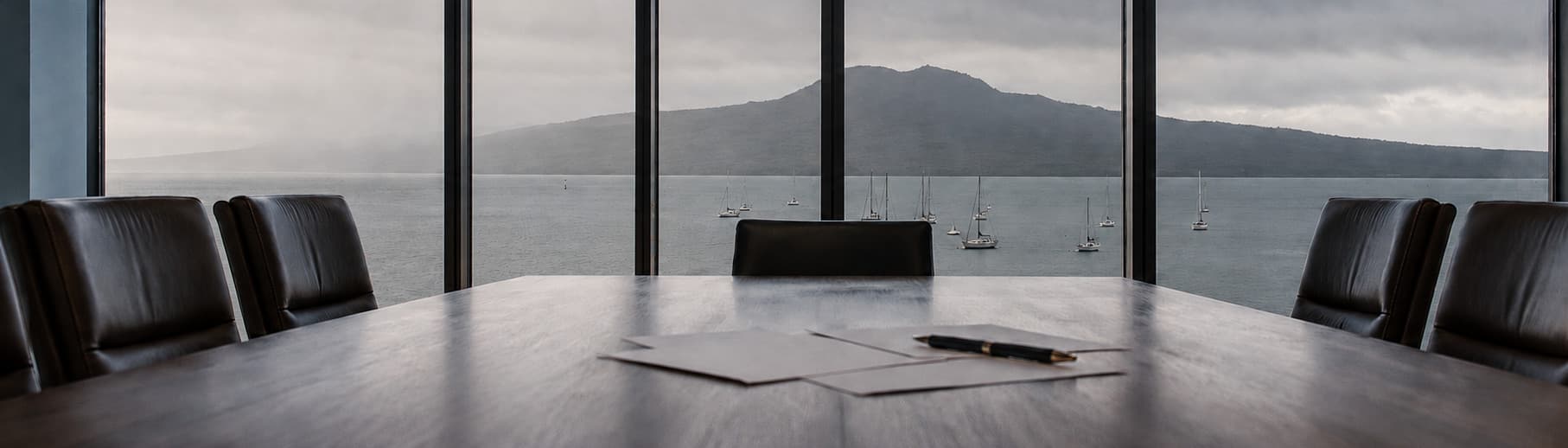 Empty meeting room table with papers and a pen, windows looking across the harbour to Rangitoto Island under heavy cloud