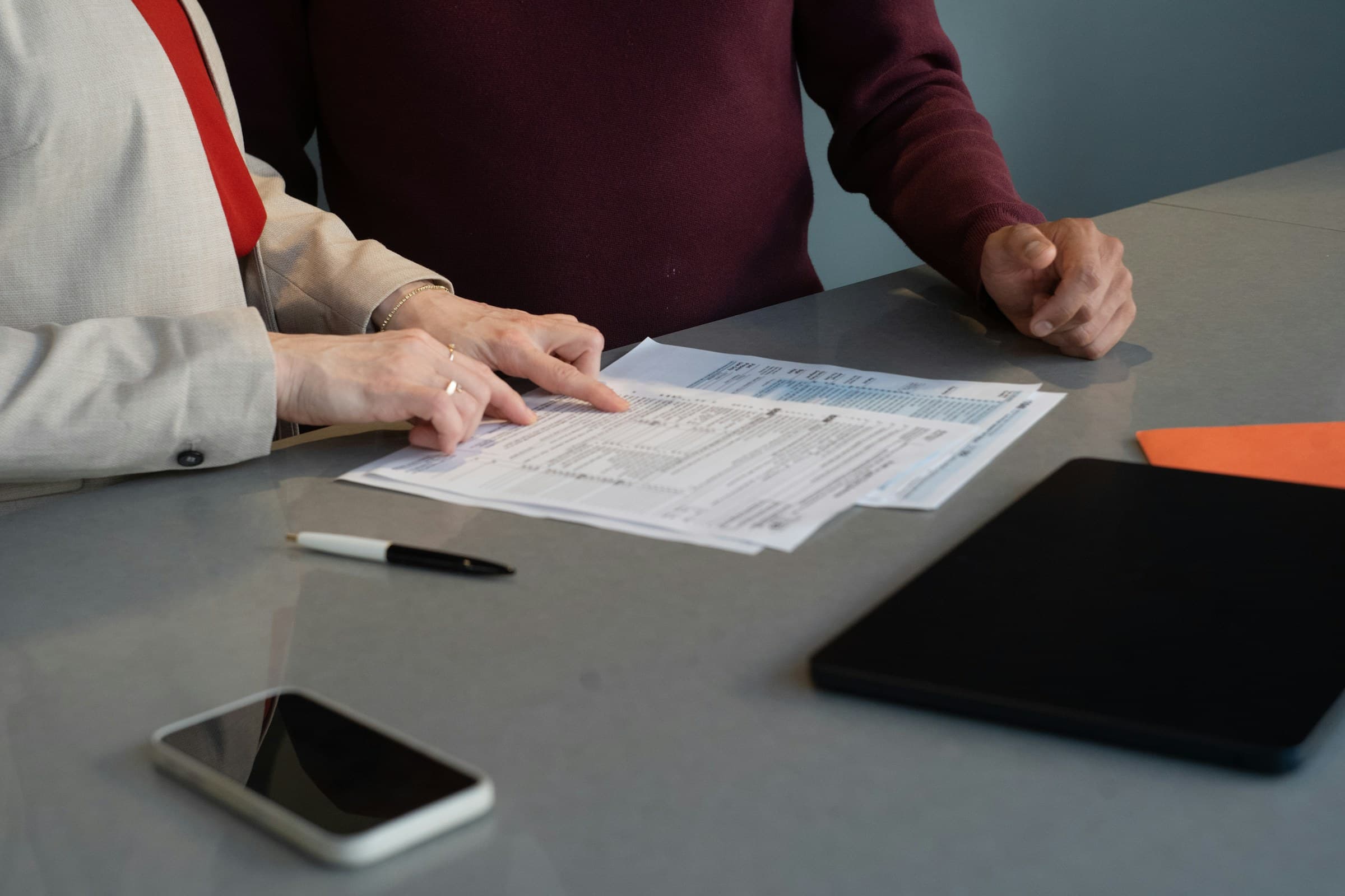 One person pointing to a line on a printed document while another listens, hands resting on the table